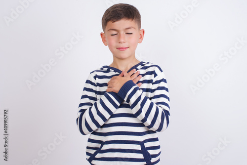little cute boy kid wearing stripped t-shirt closes eyes and keeps hands on chest near heart, expresses sincere emotions, being kind hearted and honest. Body language and real feelings concept.