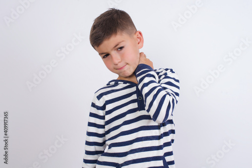 little cute boy kid wearing red stripped t-shirt against white wall suffering from back and neck ache injury, touching neck with hand, muscular pain.