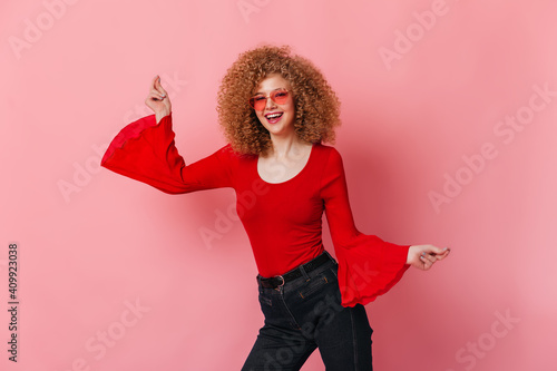 Positive girl in red top with wide sleeves and colored glasses dancing on pink background