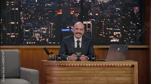 Late-night talk show host sitting behind his table and performing his monologue, looking into camera. TV broadcast style show