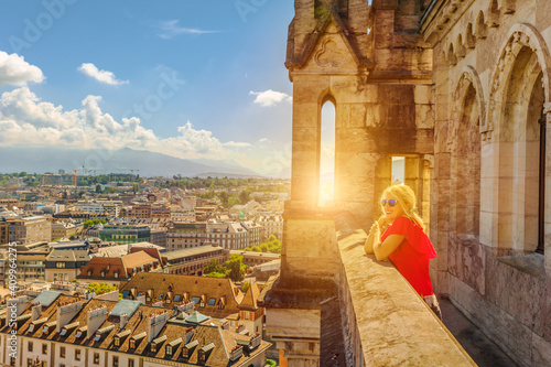 Woman tourist enjoys view over rooftops of Geneva skyline from height of the north tower of Cathedral Gothic Romanesque of Saint-Pierre, French Swiss in Switzerland. Sunset light between mountains.