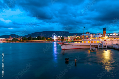 Dramatic stormy sky of Geneva urban cityscape and Swiss Alps reflecting in Lake Leman, Switzerland in French Swiss. Pier in Quai du Mont-Blanc ferry terminal at dusk, illuminated in the evening.