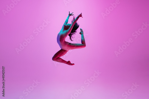 High above. Young and graceful ballet dancer on pink studio background in neon light. Art, motion, action, flexibility, inspiration concept. Flexible caucasian ballet dancer, moves in glow.