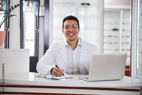 Smiling bespectacled optometrist seated at the table in his office