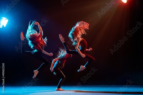 group of three ballet girls in tight-fitting costumes jumping on black background with their long hair down, silhouettes illuminated by color sources