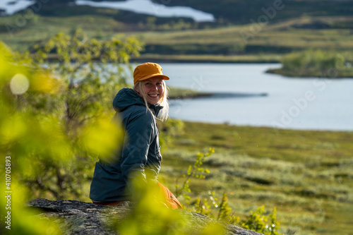 Caucasian outdoor active young adult woman sitting on Rock With outdoor hiking cloths jacket and cap smiling and looking at camera with montain and lake background in Padjelanta national park, Sweden.