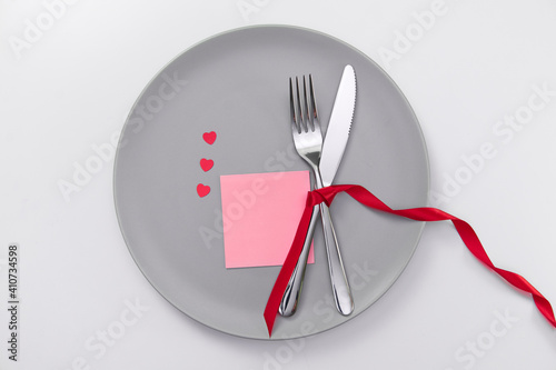 Valentine's day festive table setting, flat lay with red heart shape on gray plate, fork, knife and red ribbons on white table. Love dating concept. Place your text and copy space. High quality photo