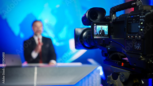 Professional TV Camera Standing in Live News Studio with Anchor seen in Small Display. Unfocused TV Broadcasting Channel with Presenter, Newscaster Talking. Mock-up Television Channel Newsroom Set