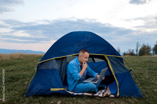 Man working on laptop in tent in nature. Young freelancer sitting in camp. Relaxing in mountains. Remote work, outdoor activity in summer. Happy male work on vacation.