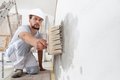 construction worker painter man with protective helmet, brush in hand restores and paint the wall, inside the building site of a house, closeup