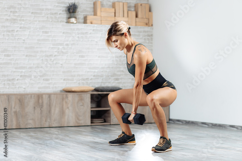 Strong young blonde woman doing heavy dumbbell exercise in gym. Female doing squats.