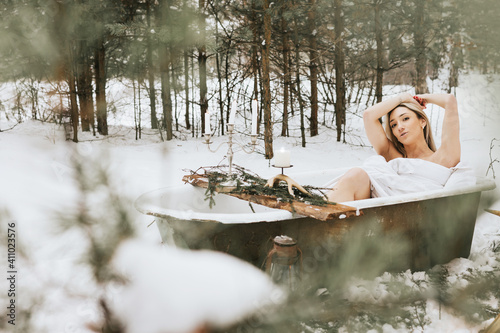 young beautiful blonde woman sits and enjoying in a cast iron bath in a snowy forest with candles and a bonfire, the concept of creative fashion and unusual leisure