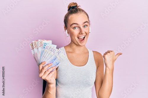 Beautiful blonde sport woman holding 20 swedish krona banknotes pointing thumb up to the side smiling happy with open mouth