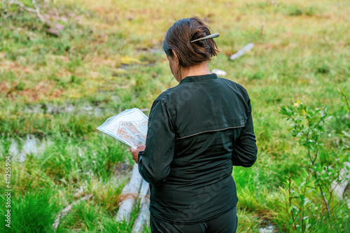 Cute middle aged Caucasian women wearing black sportswear standing at marsh edge and looking at map during exercise in outdoor orienteering in pine tree forest, Sweden, hobby sport, back side view