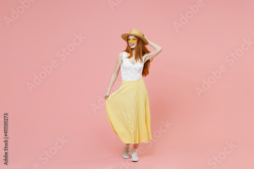 Full length body nice young fashionable redhead ginger stylish woman in straw hat glasses summer clothes yellow pleated skirt looking camera isolated on pastel pink color background studio portrait.
