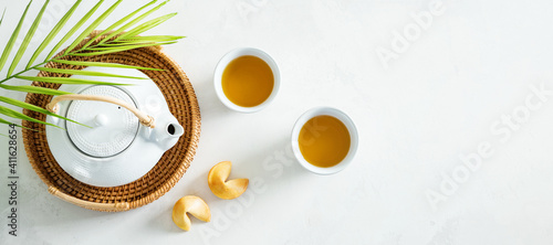 View from above of Asian tea concept, two white cups of tea, fortune cookies and teapot surrounded with dry green tea on light background.Wide banner.