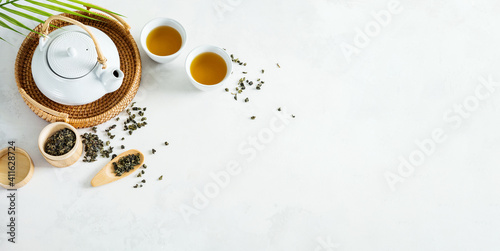Asian tea concept, two white cups of tea and teapot surrounded with green dry tea with space for a text on light background. Wide banner.