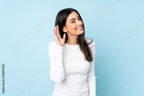 Young caucasian woman isolated on blue background listening to something by putting hand on the ear