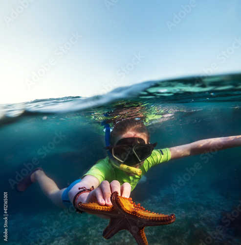 Underwater shot of a young teenage boy snorkeling with dive face mask in the blue Indian Ocean waves near the Zanzibar island with a big orange starfish in his hand. Exotic vacations concept image.