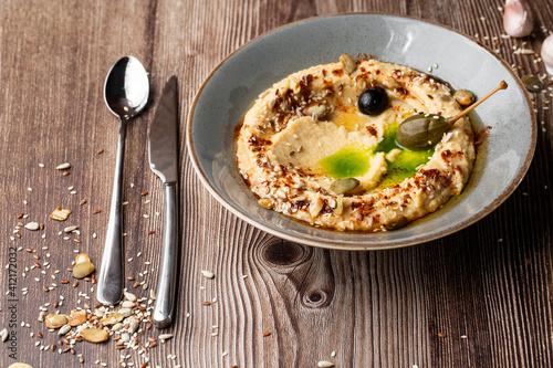 Hummus in a grey bowl seasoned with sunflower and pumpkin seeds, sesame, olives and olive oil. Close-up on a table with cutlery, seeds and garlic by side. Isolated dish on wooden background.