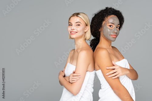 Multicultural women in facial masks and towels looking at camera isolated on grey