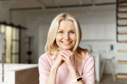 Portrait of a charming elegant mature middle-aged businesswoman with hands on a chin smiling at the camera, sitting in the modern house, wearing pink sweater. The view while the video-calling concept