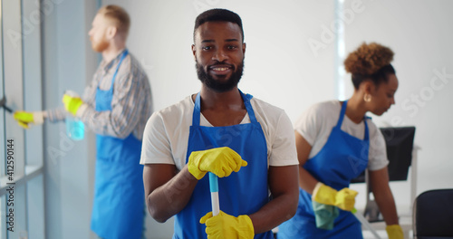 Portrait of african cleaner in apron and gloves holding mop and smiling at camera working in office