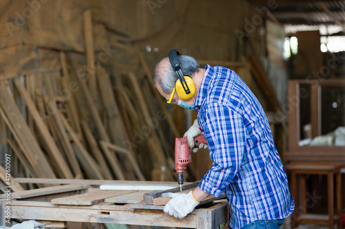 Asian senior male carpenter using electric drill on wood and wearing safety goggles and earmuffs at work..Asian woodworker working at woodwork workshop. Asian workman carpenter