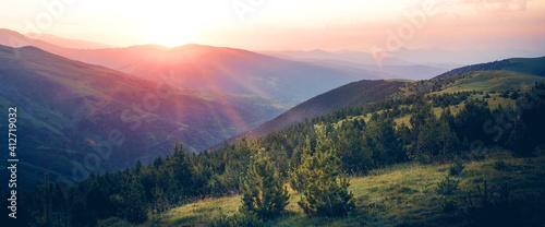 Beautiful sunrise on top of the mountain with fir trees. A new day begins. Sunbeams in the golden hour. Concept of nature, holidays, and healthy lifestyle. Panoramic landscape view.
