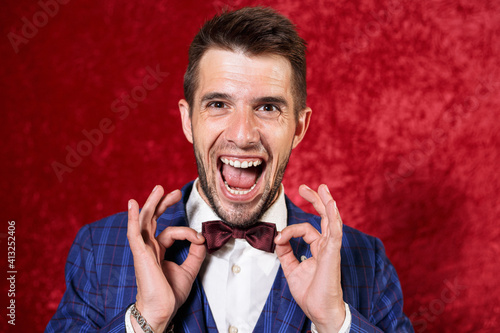 Cheerful charismatic showman in suit touching bow tie while standing on red background in stud and looking at camera during eccentric show 