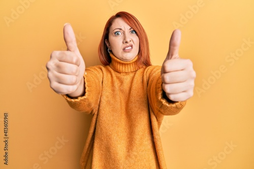 Beautiful redhead woman doing thumbs up positive gesture in shock face, looking skeptical and sarcastic, surprised with open mouth