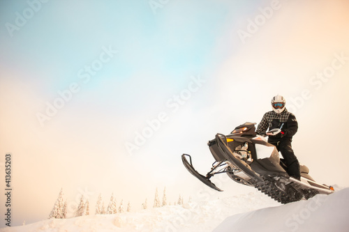 The rider in gear with a helmet making snow jumping on a snowmobile on a background of a winter scenic landscape with mounting and sky.