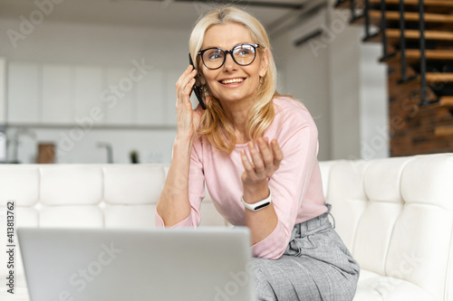 Beautiful positive middle-aged blond woman smiling and chatting on the phone, sitting at white couch at home,talking with coleagues,working at home,explaining,making a meeting