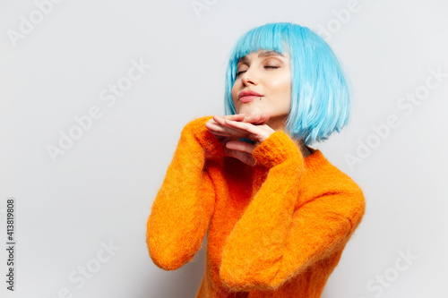 Portrait of young girl with blue hair in orange sweater, holding hands under chin against white background.
