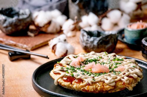 Homemade japanese fast food okonomiyaki cabbage pancake with onion, pickled ginger, mayo sauce on black ceramic plate. Chopsticks, teapot, cotton, ingredients above. Texture background. Flat lay
