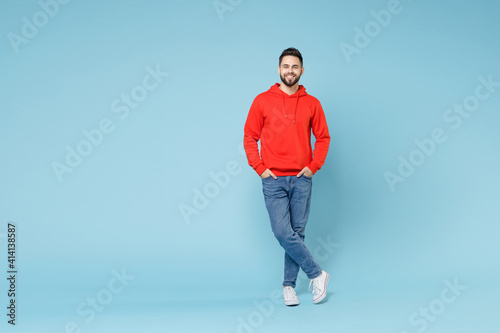 Full length young caucasian smiling bearded handsome student man 20s wear casual red orange hoodie standing akimbo arms on waist isolated on blue background studio portrait People lifestyle concept