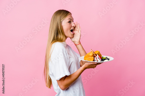 Young russian woman eating a waffle isolated shouting and holding palm near opened mouth.