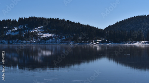 Peaceful view of frozen lake Schluchsee in the Black Forest hills, Germany in winter season with coniferous forest reflected on the ice surface on sunny day with blue sky.