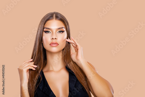 Beauty Portrait Young Woman holding her long natural brown Hair with her hands. Makeup. Brunette model with long hair posing over Beige background. Girl looking at camera. Eye makeup, peach lips 