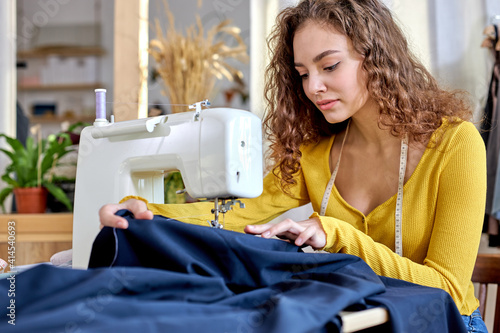 young tailor working on sewing machine, hand making clothes in home interior. caucasian curly female sits enjoying process of sewing clothes. creative skills design, hobbies and lifestyle concept