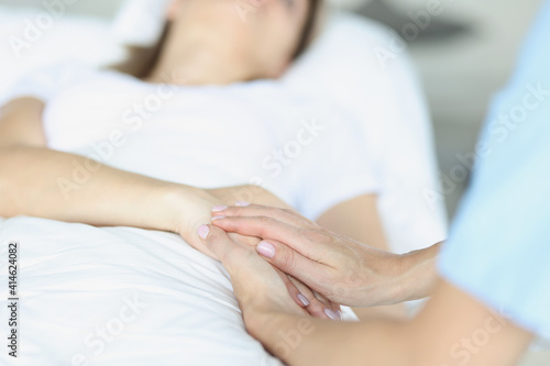 On bed lies a sick doctor sitting next to him and sympathetically holds hand