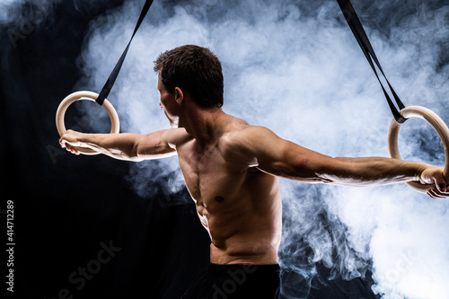 Muscular build man doing calisthenics on gymnastics rings indoor on black, smoked background. concept of healthy lifestyle and power.