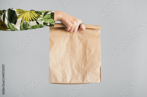 Female hand holds a paper bag against gray backdrop. Empty eco friendly package, gift, surprise or delivery concept