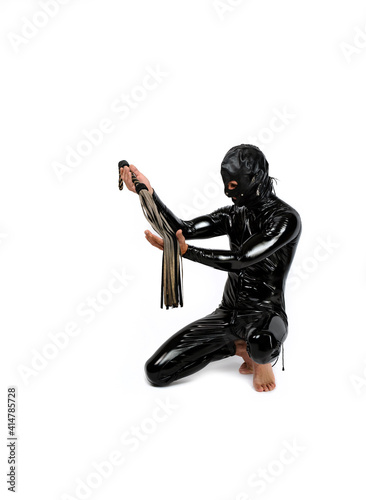 Slave sitting on his knees. Man dressed in latex rubber fetish clothes and pvc mask. Slave bowed his knees and holds a leather whip in hands. bdsm costume for role-playing adult games.