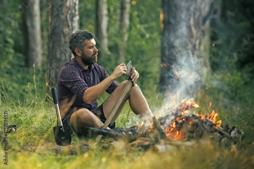 Man hipster sharpen axe. Camping, hiking, summer vacation. Guy relax at bonfire in forest.