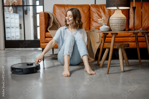 Black robotic vacuum cleaner cleaning the floor while woman sitting near sofa and relax. Smart technology concept. High quality photo