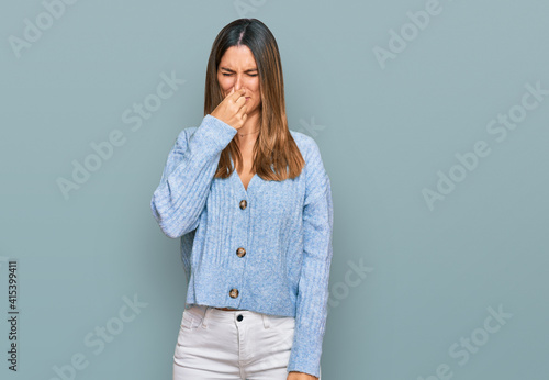 Young woman wearing casual clothes smelling something stinky and disgusting, intolerable smell, holding breath with fingers on nose. bad smell