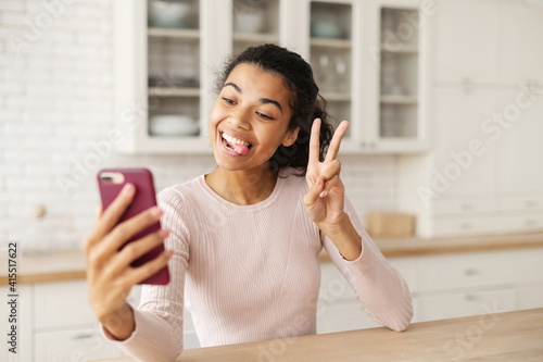 Funny biracial teen with dark hair and bronze skin showing tongue and taking photo on the phone, making a selfie, showing a piece sign, at home in the kitchen by the tabletop, chatting with friends