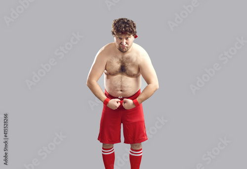 Funny topless strong plump athlete in red gym shorts showing his chubby body standing isolated on gray background. Young man with curly hair and some belly fat flexing arms pretending to be tough guy