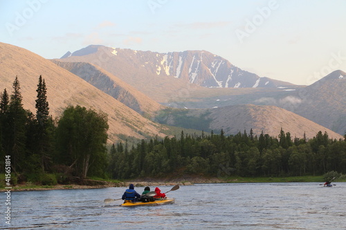 Beautiful mountain landscape with snow streaks on the slopes illuminated by the setting sun with a forest on the lake shore and a tourist kayak with people on the water.Smooth hills lit up in the haze